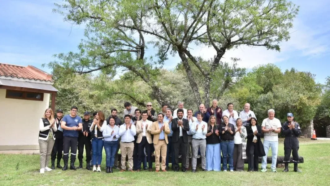 Unas treinta personas, entre funcionarios de civil y personal uniformado de la Policía y de organismos de emergencia, posan aplaudiendo sobre el césped de un predio arbolado, junto a una construcción de techo de tejas.