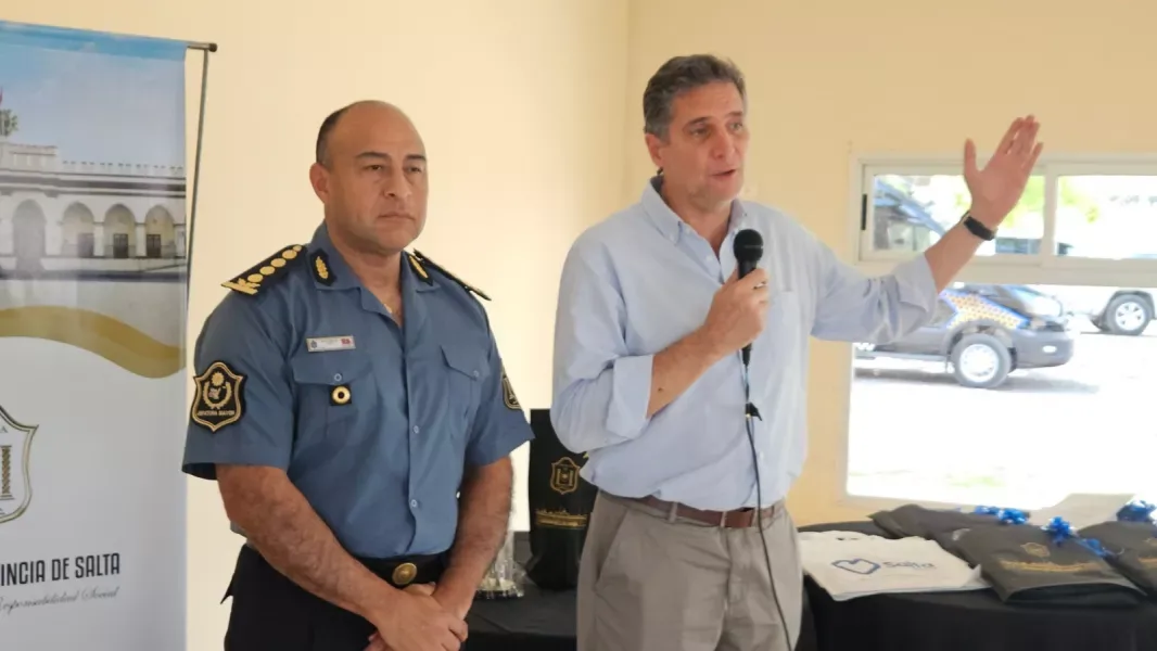 Un hombre de camisa celeste habla con un micrófono y el brazo en alto, junto a un oficial superior uniformado de la Policía de Salta, delante de un banner del Gobierno de la Provincia; sobre una mesa hay bolsas y presentes y por la ventana se ve un móvil policial.