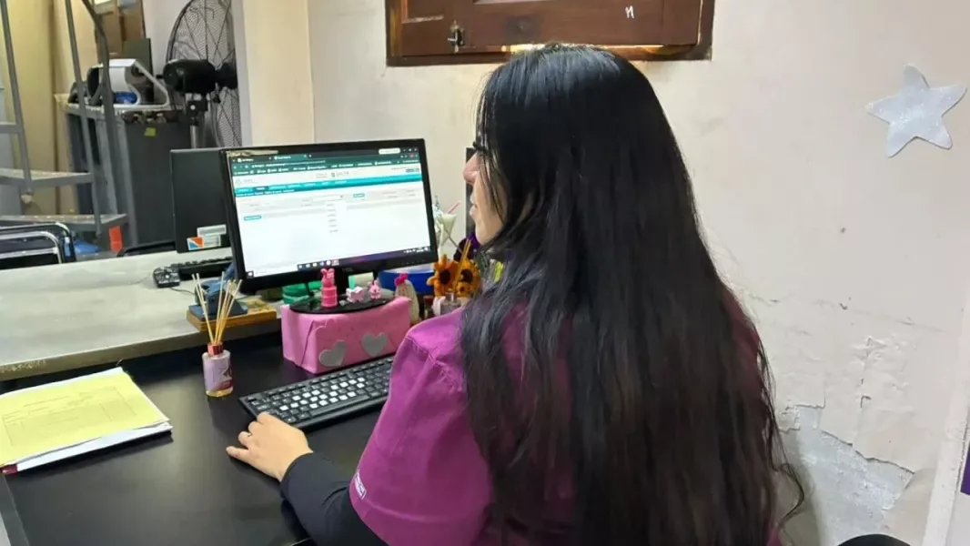 Una trabajadora de salud, de espaldas, carga datos en una computadora de escritorio en una oficina de turnos hospitalaria