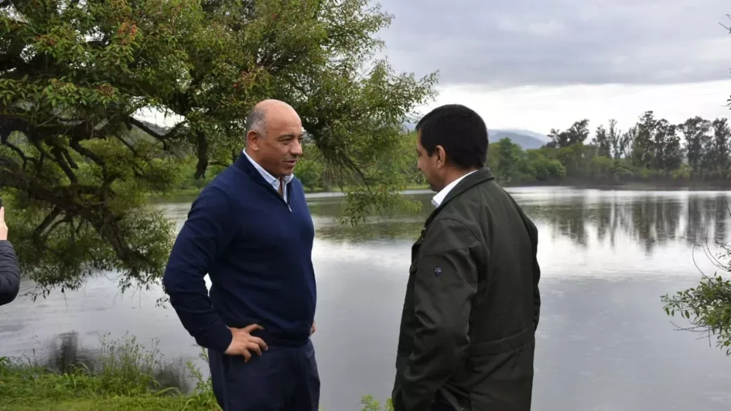 Dos hombres conversan de pie a la orilla del dique de Rosario de la Frontera, con el agua y los árboles detrás y el cielo nublado