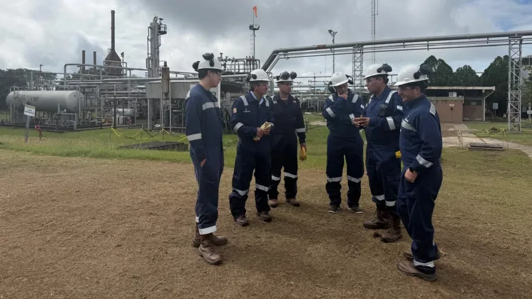 Seis hombres con mamelucos azules y cascos blancos conversan en un descampado frente a una planta de tratamiento de gas, con torres, cañerías y tanques rodeados de vegetación.