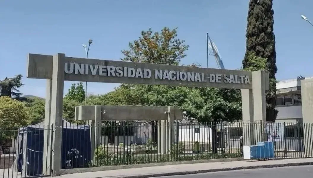 Portón de acceso a la Universidad Nacional de Salta sobre avenida Bolivia, con el cartel de la universidad y la vereda frente a la reja