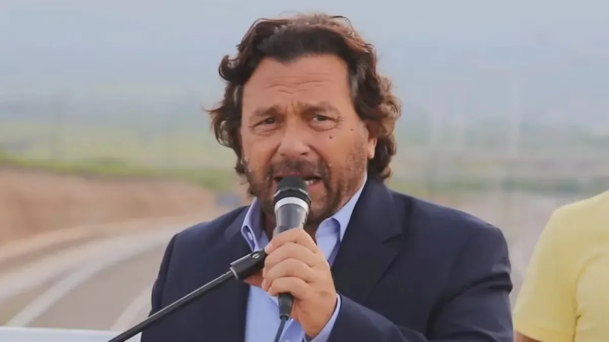 El Gobernador de Salta, Gustavo Sáenz, de pelo ondulado y barba, con saco azul y camisa celeste, habla frente a un micrófono al aire libre, con una ruta y cerros borrosos de fondo.