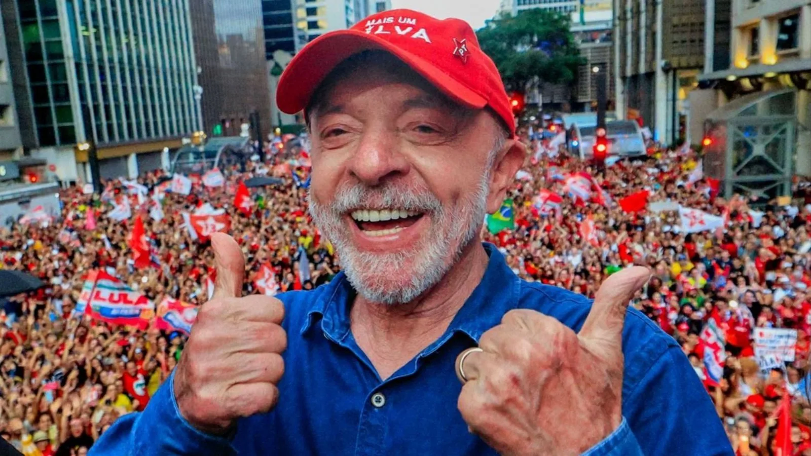 Un hombre mayor de barba blanca, con gorra roja y camisa azul, sonríe con los pulgares en alto frente a una multitud con banderas rojas en una avenida.
