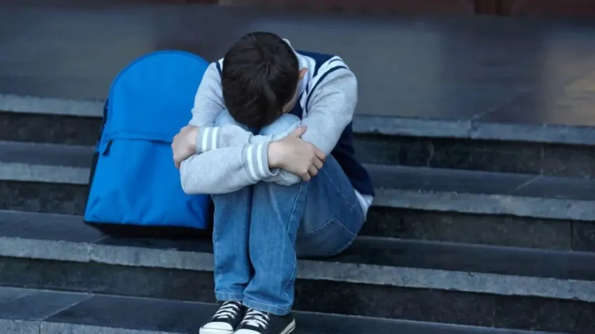 Un chico sentado en la escalera de una escuela, con la cabeza escondida entre las rodillas y la mochila al lado