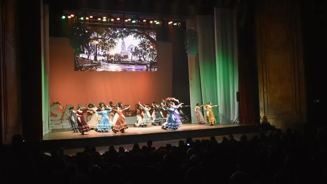 Bailarinas con polleras de colores sobre el escenario del Teatro Provincial, con la imagen de la Catedral de Salta proyectada al fondo