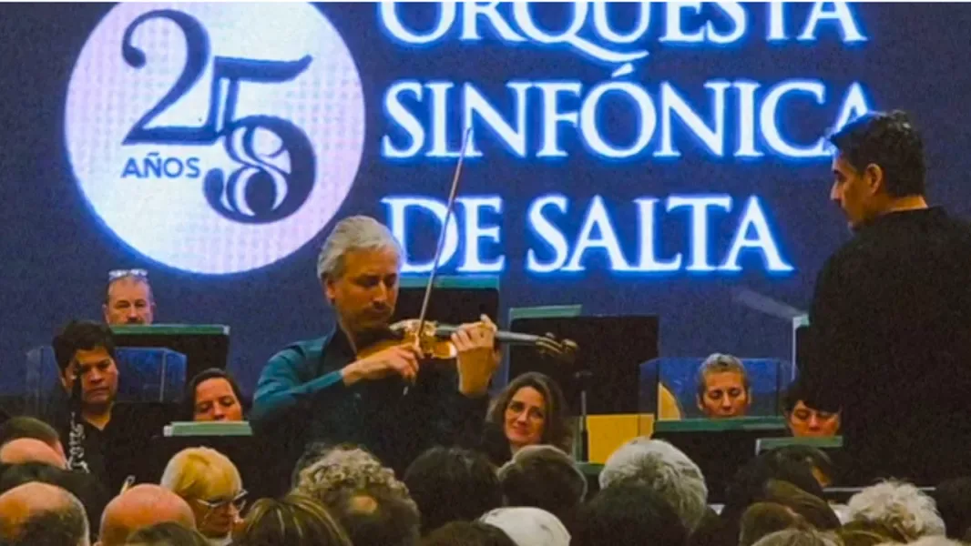 Un violinista solista toca frente al público junto al director, delante de una pantalla con el logo de los 25 años de la Orquesta Sinfónica de Salta