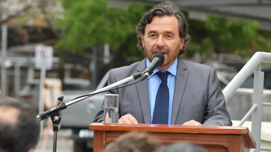El gobernador Gustavo Sáenz habla ante un micrófono en un atril de madera, al aire libre, con árboles de fondo y público en primer plano