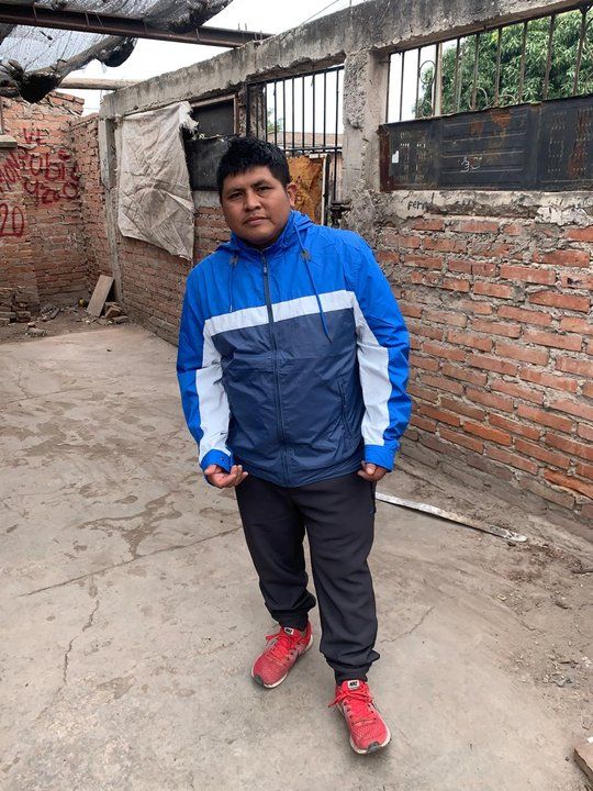 Hombre joven de contextura media y cabello negro corto, de pie en un patio, con campera azul y blanca, pantalón de jogging oscuro y zapatillas rojas