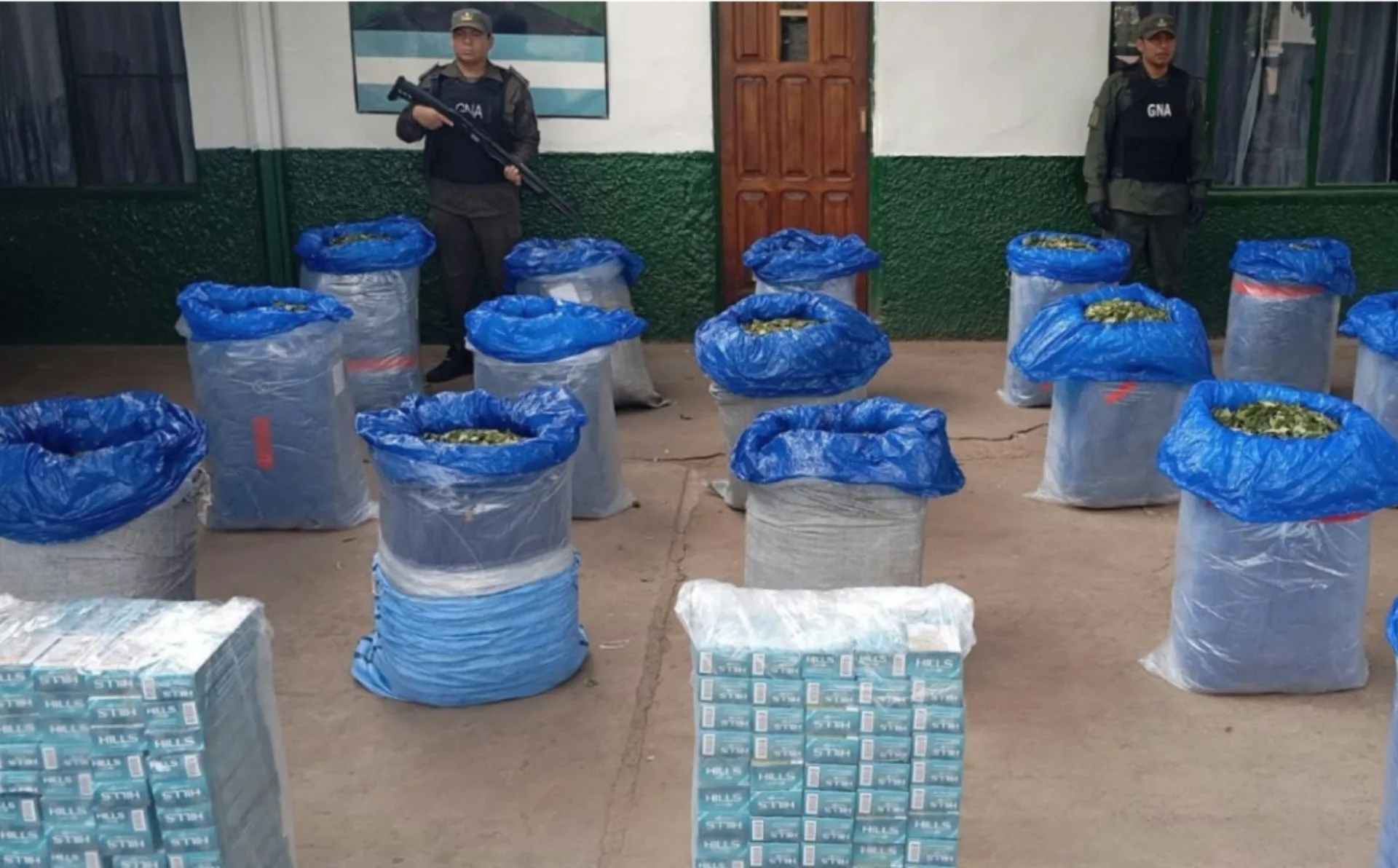 Bolsas de hojas de coca y paquetes de cigarrillos decomisados, con dos gendarmes custodiando frente a un edificio de Gendarmería Nacional