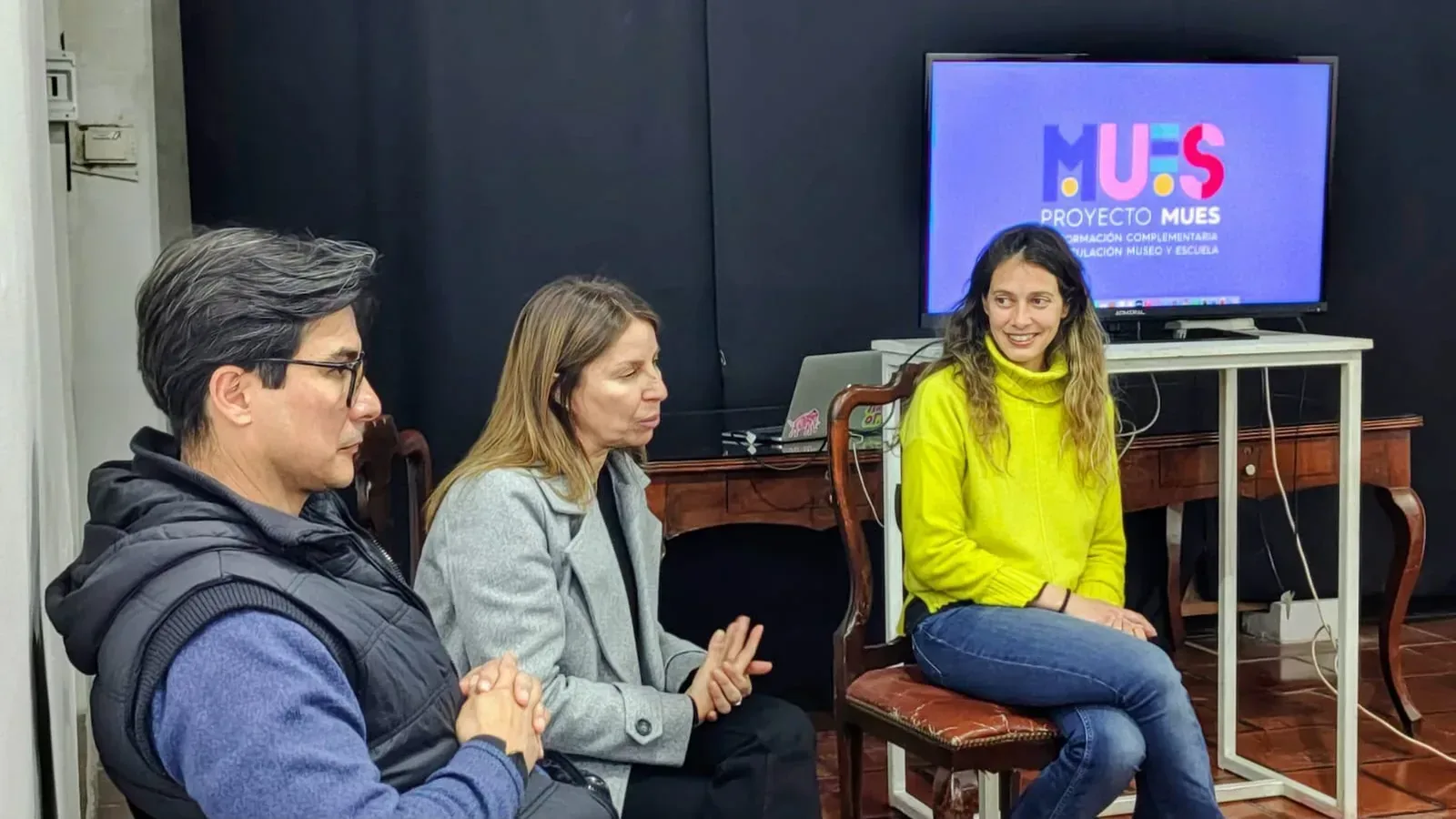 Tres personas conversan frente a una pantalla con el logo del proyecto MUES durante la clase de formación