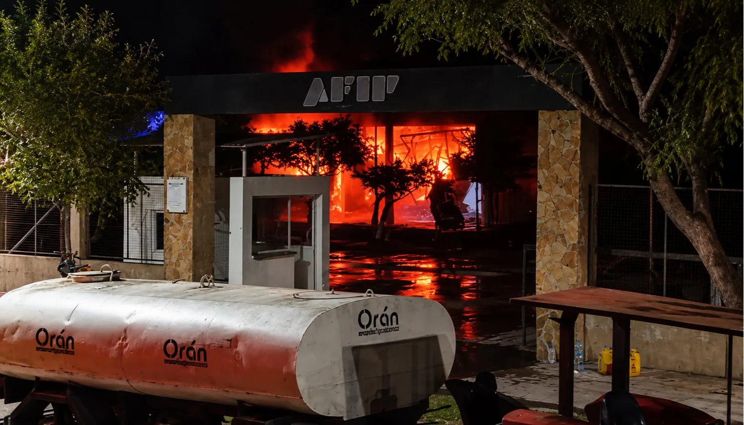 El fuego arde detrás del portón de acceso con el cartel de AFIP del predio de la Aduana en Orán, de noche, con una cisterna municipal de agua en primer plano