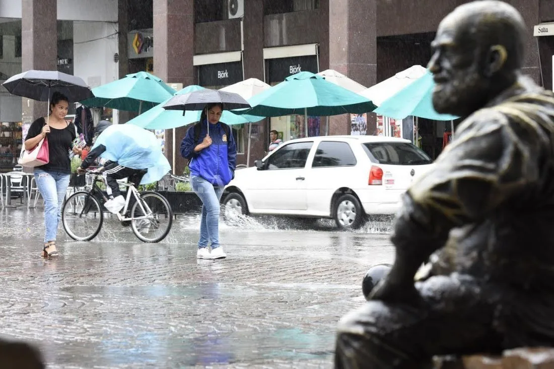 Peatones con paraguas cruzan una calle mojada del centro de Salta bajo la lluvia, junto a una escultura de bronce