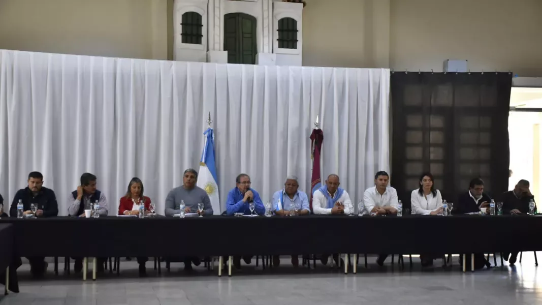 Once funcionarios, intendentes y legisladores sentados detrás de una mesa larga con mantel negro, frente a un telón blanco con la bandera argentina y la de Salta