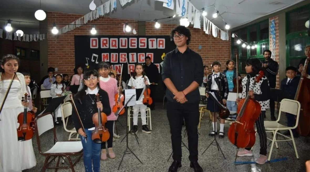 Chicos y chicas de la orquesta de la Fundación Música con Alas, con violines y violonchelos, frente al cartel de la orquesta