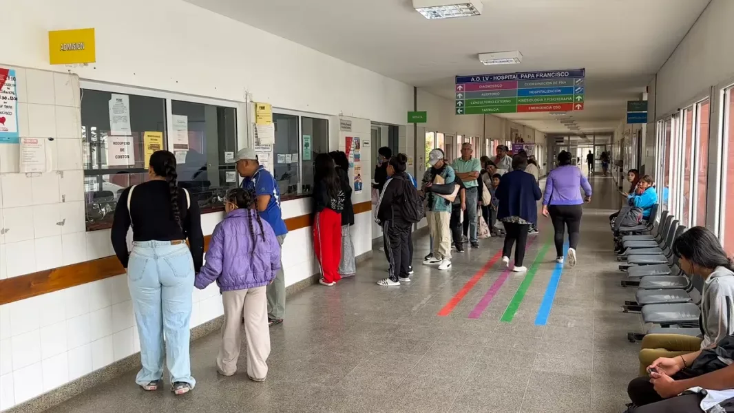 Pasillo de admisión del Hospital Papa Francisco de Salta, con personas haciendo fila frente a las ventanillas, otras sentadas en la sala de espera y líneas de colores pintadas en el piso que guían hacia los distintos servicios.