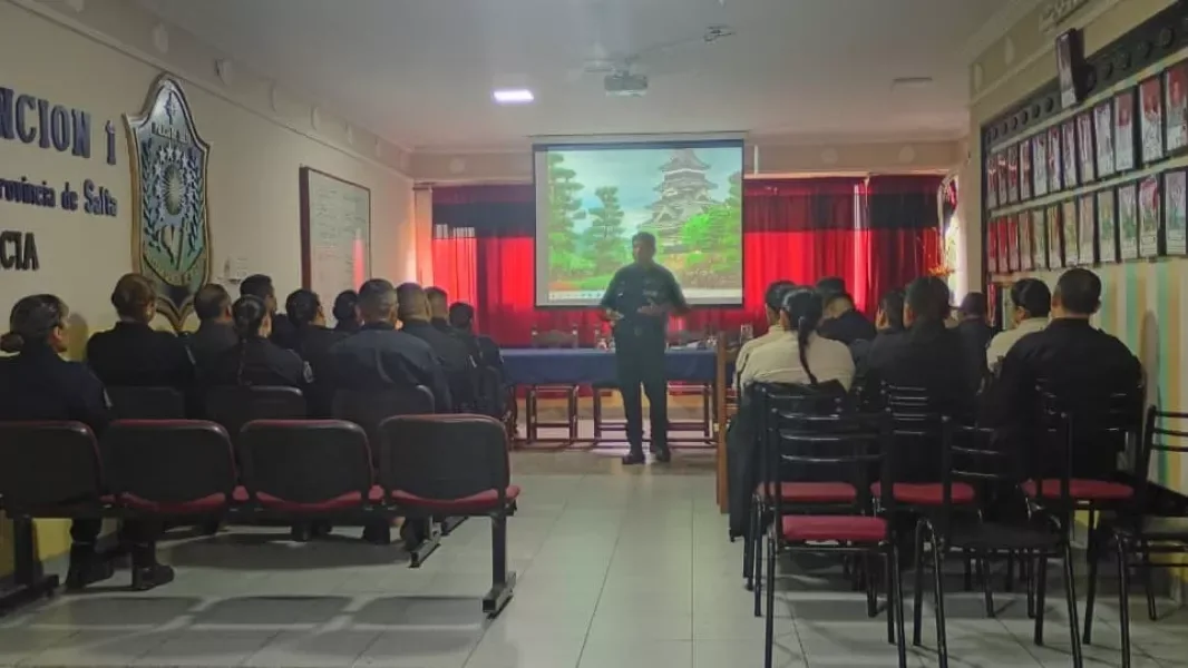Personal policial uniformado, de espaldas, sentado en un salón durante una reunión; al frente un oficial expone junto a una pantalla, bajo el escudo de la Policía de Salta