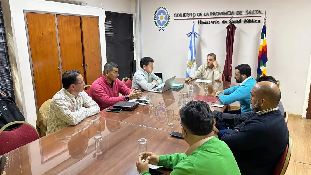 Ocho hombres sentados alrededor de una mesa larga de madera en una sala de reuniones, con el escudo del Gobierno de la Provincia de Salta y el cartel del Ministerio de Salud Pública en la pared del fondo, junto a banderas.