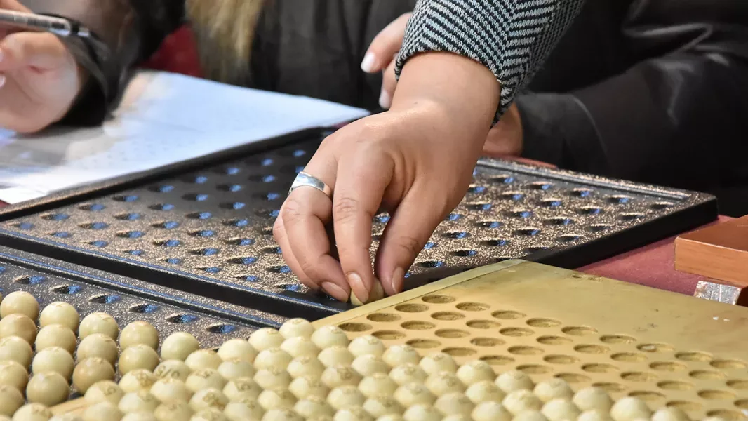 Primer plano de las manos de una mujer que acomoda bolillas numeradas en las bandejas oscuras de un bolillero. Delante hay otra bandeja de madera clara llena de bolillas.