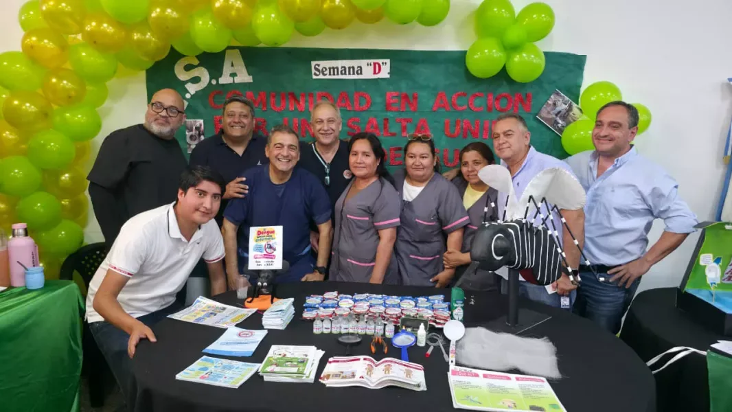 Una decena de personas, entre funcionarios y personal de salud con ambo, posan detrás de una mesa con folletos, frascos de larvicida, una lupa y un mosquito gigante de utilería. Al fondo, un cartel verde que dice Semana D y un arco de globos verdes y amarillos.