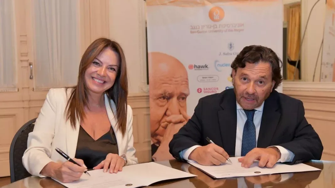 El gobernador Gustavo Sáenz y la ministra de Capital Humano, Sandra Pettovello, firman sendas carpetas sentados a una mesa. Detrás hay un banner de la Universidad Ben-Gurión del Néguev con el retrato de un hombre mayor y logos de empresas.