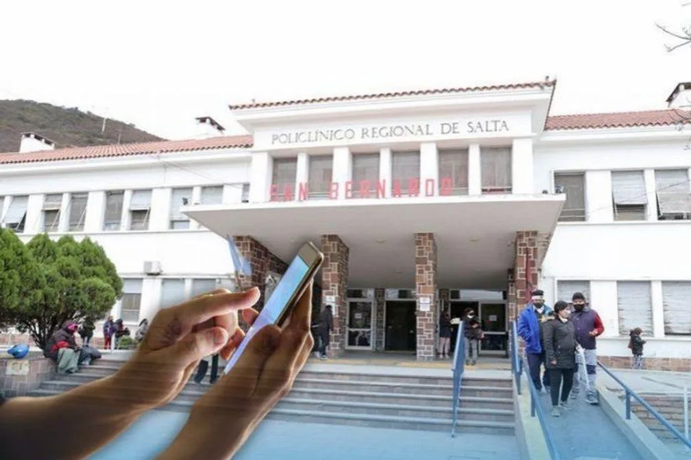 Fachada del hospital San Bernardo, en la ciudad de Salta, con una mano que sostiene un celular en primer plano