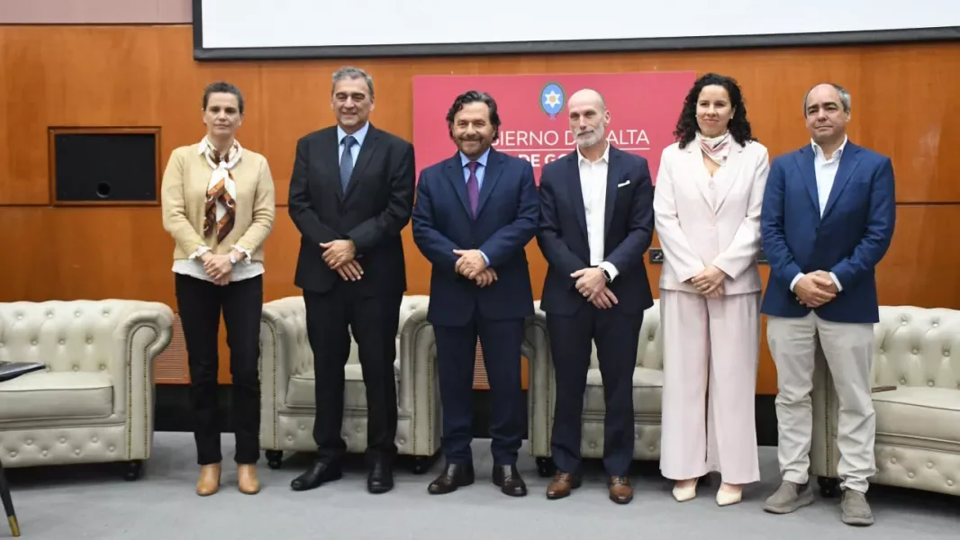 Siete personas de traje posan de pie, en fila, frente a un panel con el logo del Gobierno de Salta, en un auditorio con sillones claros detrás.