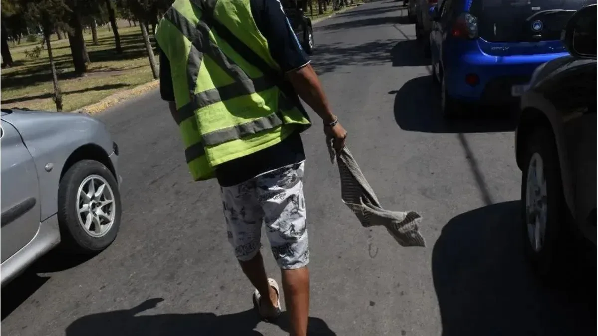 Un cuidacoches con chaleco flúor y un trapo en la mano camina entre autos estacionados