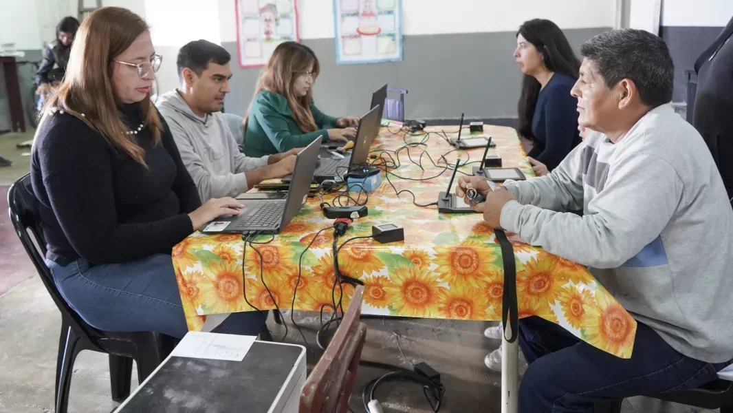 Tres empleados del Registro Civil atienden con notebooks y lectores de huellas sobre una mesa larga cubierta por un mantel de girasoles, mientras dos vecinos esperan sentados del otro lado en un salón comunitario