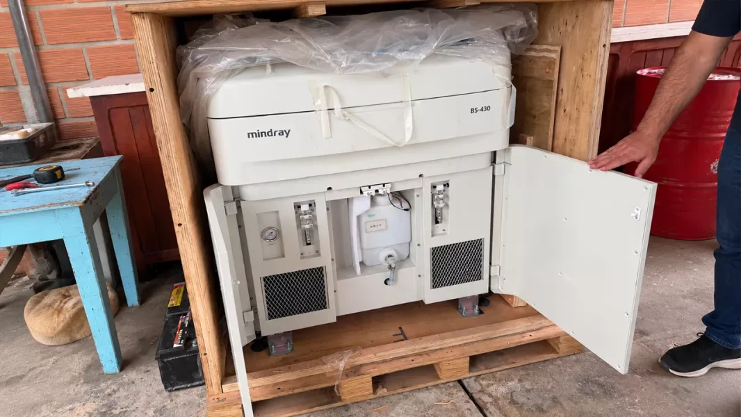 Un autoanalizador de laboratorio Mindray BS-430, de color beige, todavía dentro de su caja de madera sobre un pallet, con el embalaje plástico en la parte superior y la puerta lateral abierta por una persona de la que se ve un brazo.